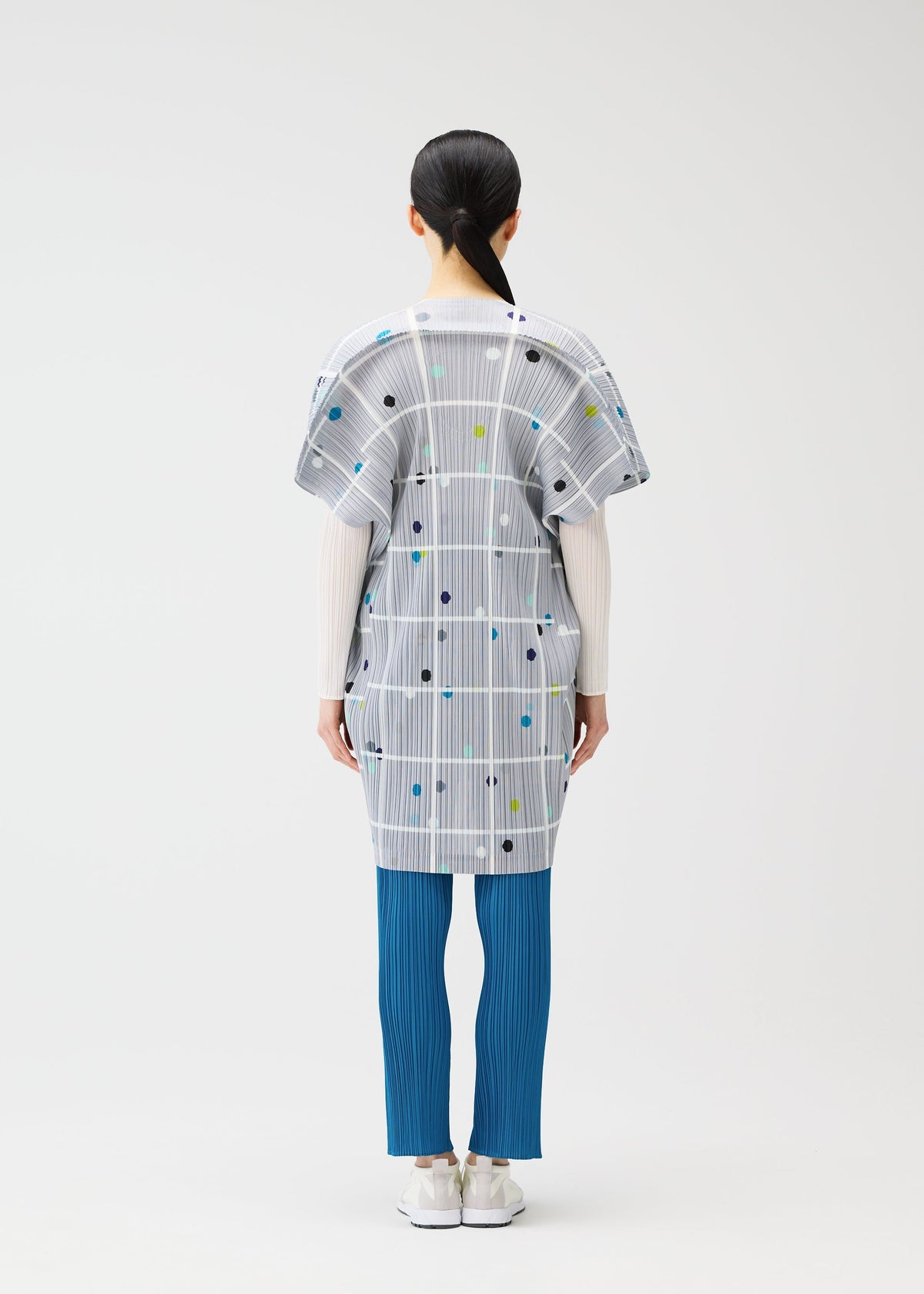 A person stands facing away, wearing the CROSSING DOTS tunic by PLEATS PLEASE ISSEY MIYAKE with city print, plaid, dots, white long sleeves, blue pleated pants, and white shoes against a plain light background. view 8 of 9