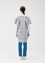A person stands facing away, wearing the CROSSING DOTS tunic by PLEATS PLEASE ISSEY MIYAKE with city print, plaid, dots, white long sleeves, blue pleated pants, and white shoes against a plain light background.