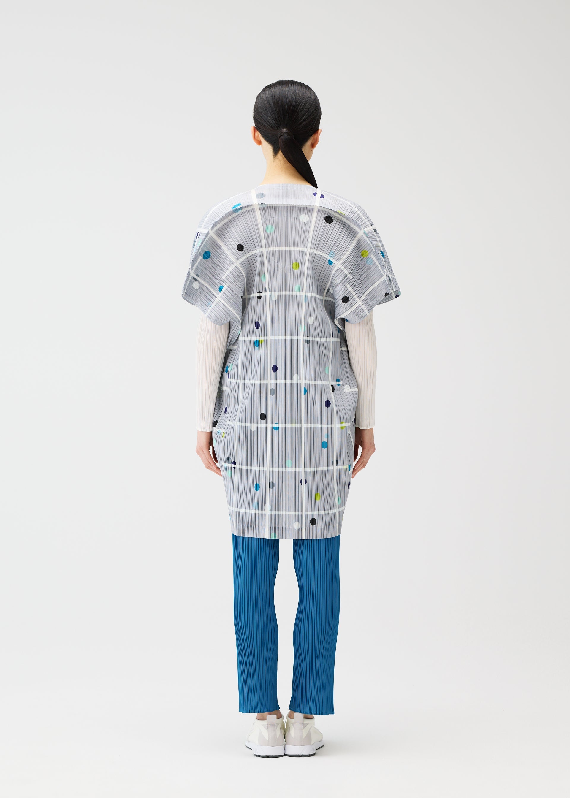 Zoomed view A person stands facing away, wearing the CROSSING DOTS tunic by PLEATS PLEASE ISSEY MIYAKE with city print, plaid, dots, white long sleeves, blue pleated pants, and white shoes against a plain light background.