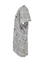 The CROSSING DOTS tunic by PLEATS PLEASE ISSEY MIYAKE is a gray, knee-length, short-sleeved piece with a round neck, featuring thin white plaid grid lines and colorful dots in black, blue, yellow, and green.
