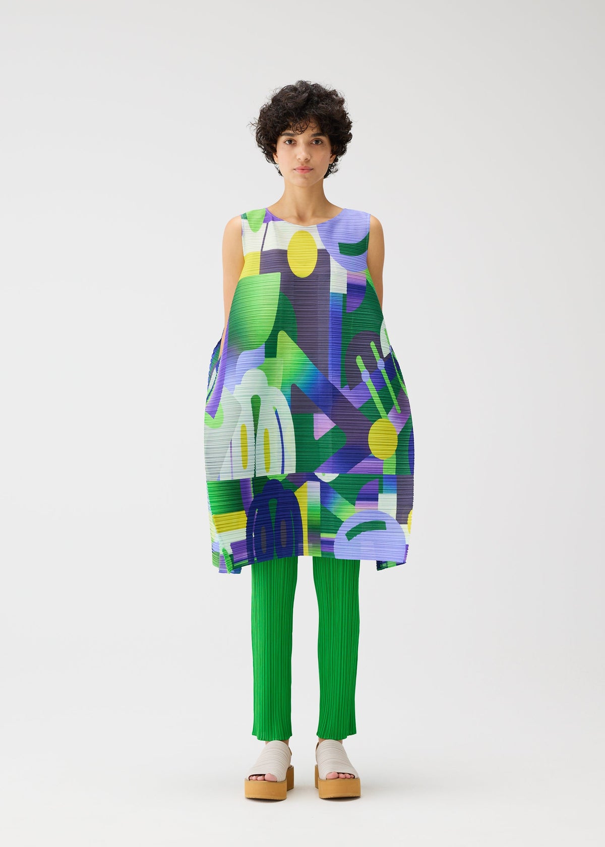 A person with short curly hair models the PLAYGROUND tunic by PLEATS PLEASE ISSEY MIYAKE—a round-neck, sleeveless piece with a colorful geometric pattern—styled over bright green pleated pants and beige platform sandals against a white background. view 6 of 9