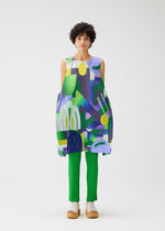 A person with short curly hair models the PLAYGROUND tunic by PLEATS PLEASE ISSEY MIYAKE—a round-neck, sleeveless piece with a colorful geometric pattern—styled over bright green pleated pants and beige platform sandals against a white background.