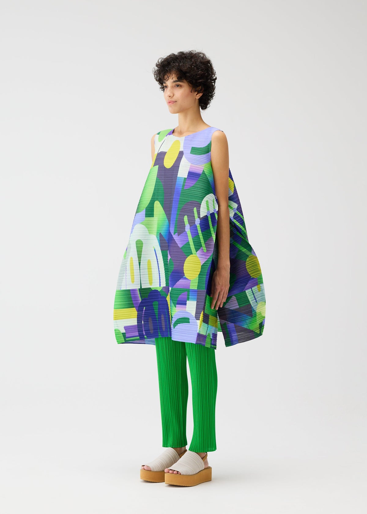 A person with short curly hair wears the PLAYGROUND tunic by PLEATS PLEASE ISSEY MIYAKE—loose, sleeveless, and colorful over bright green pleated pants and beige platform sandals—standing against a plain white background. view 7 of 9