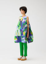 A person with short curly hair wears the PLAYGROUND tunic by PLEATS PLEASE ISSEY MIYAKE—loose, sleeveless, and colorful over bright green pleated pants and beige platform sandals—standing against a plain white background.