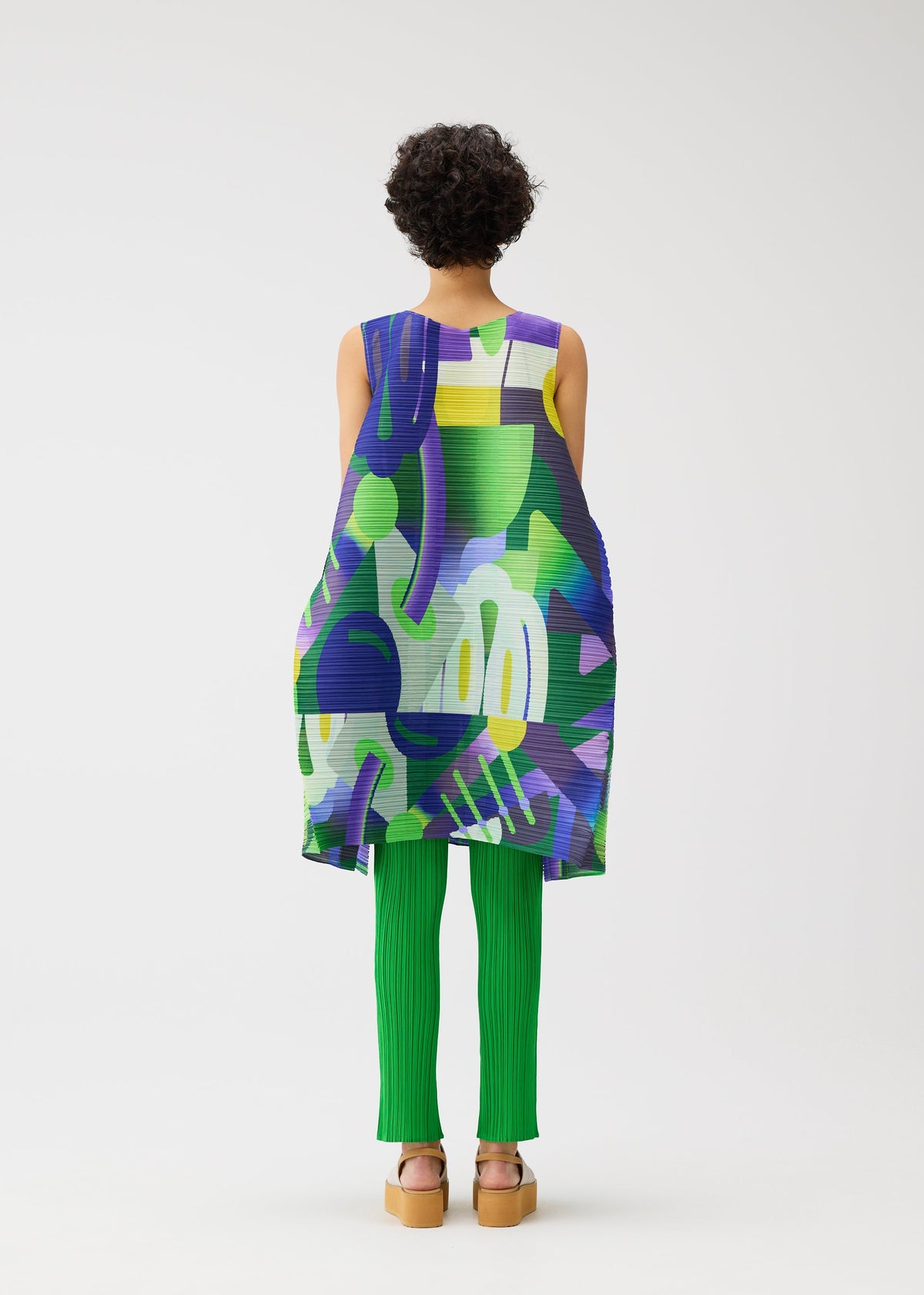A person with short curly hair, seen from behind, wears the PLAYGROUND sleeveless tunic and green ribbed pants by PLEATS PLEASE ISSEY MIYAKE, with tan platform sandals, standing against a white background. view 8 of 9