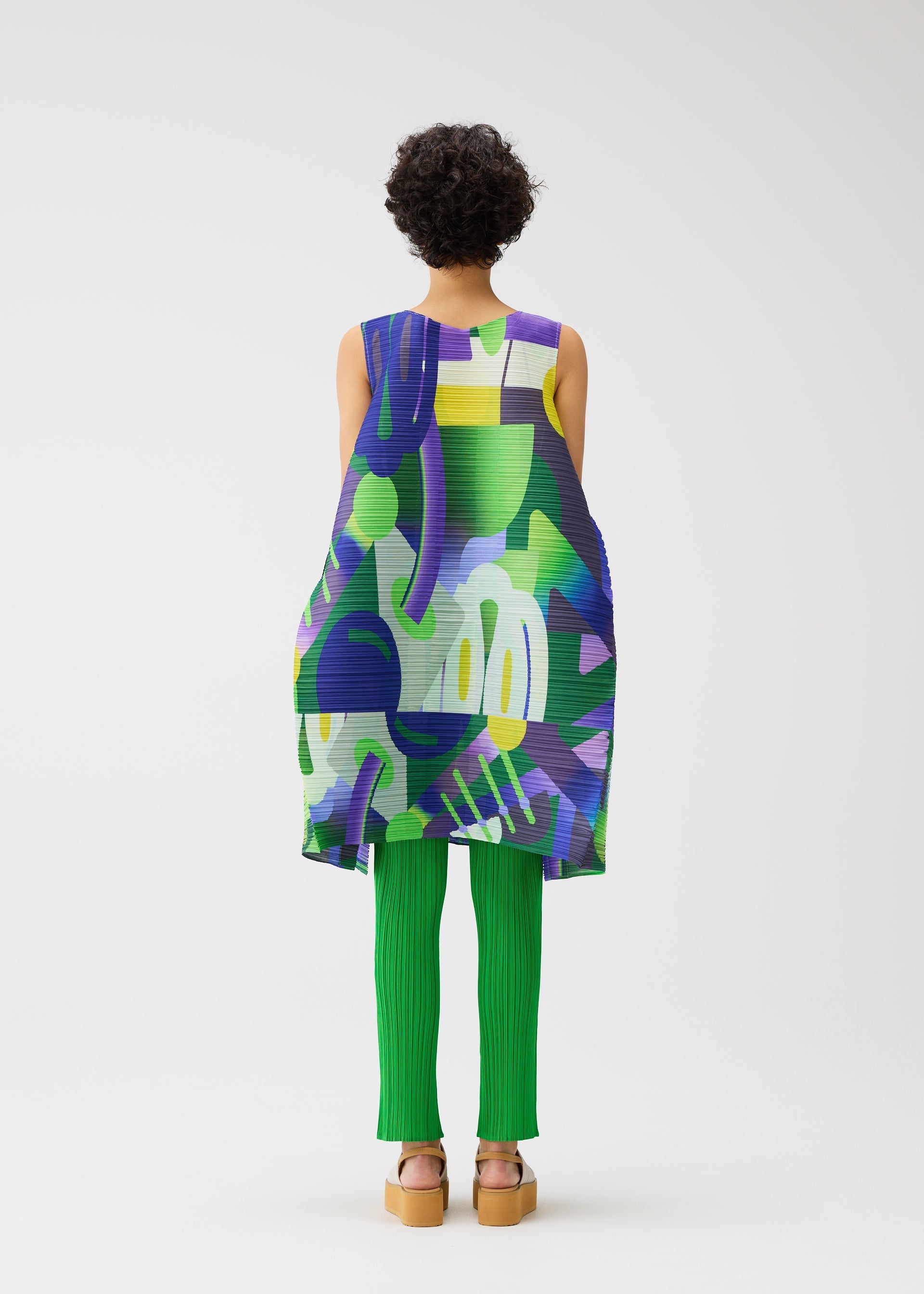 Zoomed view A person with short curly hair, seen from behind, wears the PLAYGROUND sleeveless tunic and green ribbed pants by PLEATS PLEASE ISSEY MIYAKE, with tan platform sandals, standing against a white background.