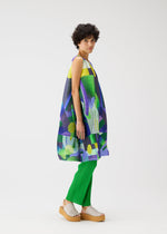 A person with short curly hair models the PLAYGROUND tunic by PLEATS PLEASE ISSEY MIYAKE—an abstract, colorful sleeveless top in purple, green, and yellow—over bright green pants and beige platform sandals against a white backdrop.