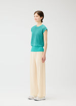 A person stands in a minimal, white space wearing the turquoise FISHNET KNIT vest by PLEATS PLEASE ISSEY MIYAKE, cream pleated wide-leg pants, and white shoes. Made from recycled nylon, the fresh look is completed with a gentle smile.