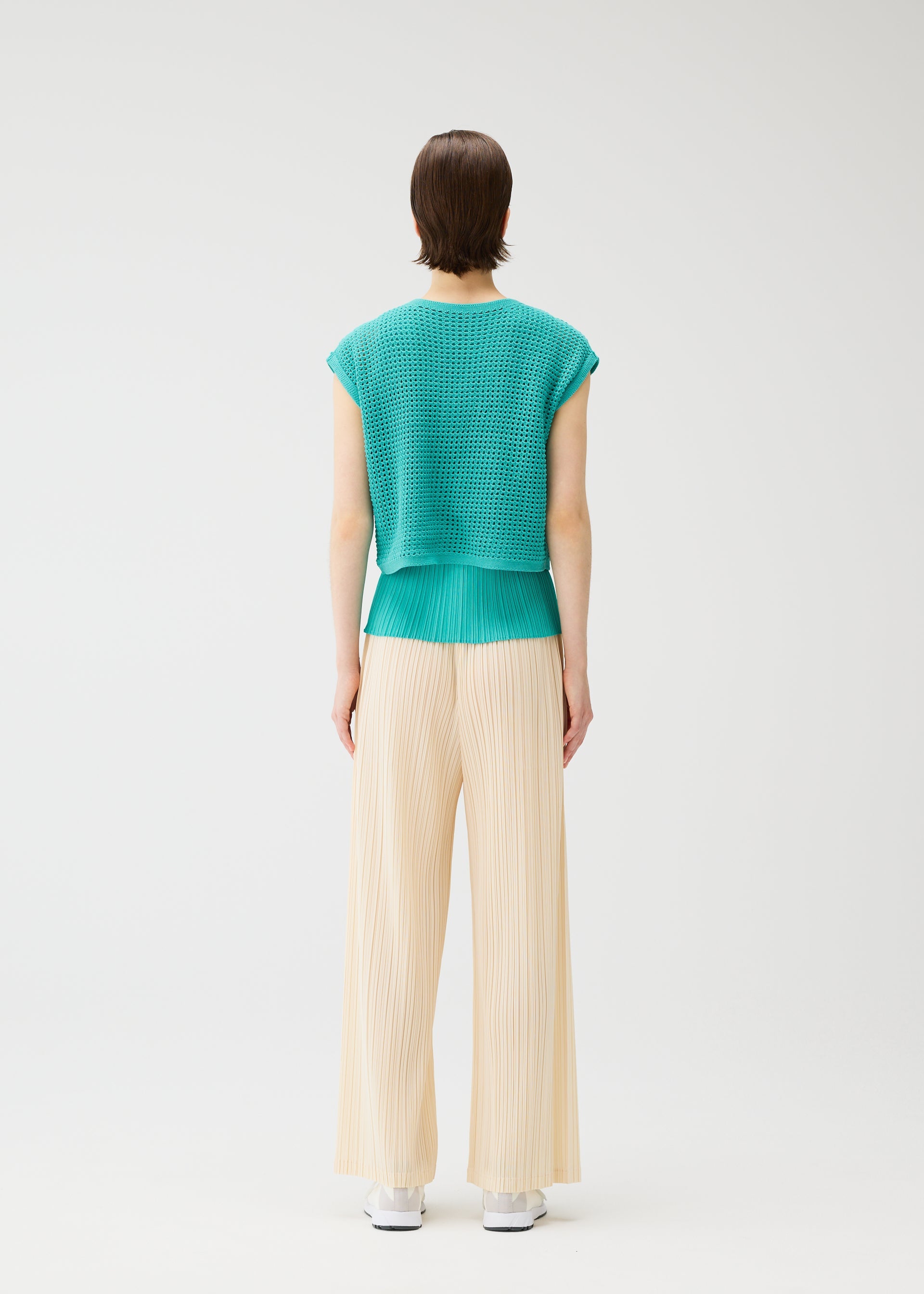 Zoomed view A person with short, straight hair faces away, wearing the PLEATS PLEASE ISSEY MIYAKE FISHNET KNIT top in turquoise and cream wide-legged pleated pants. The background is plain white.