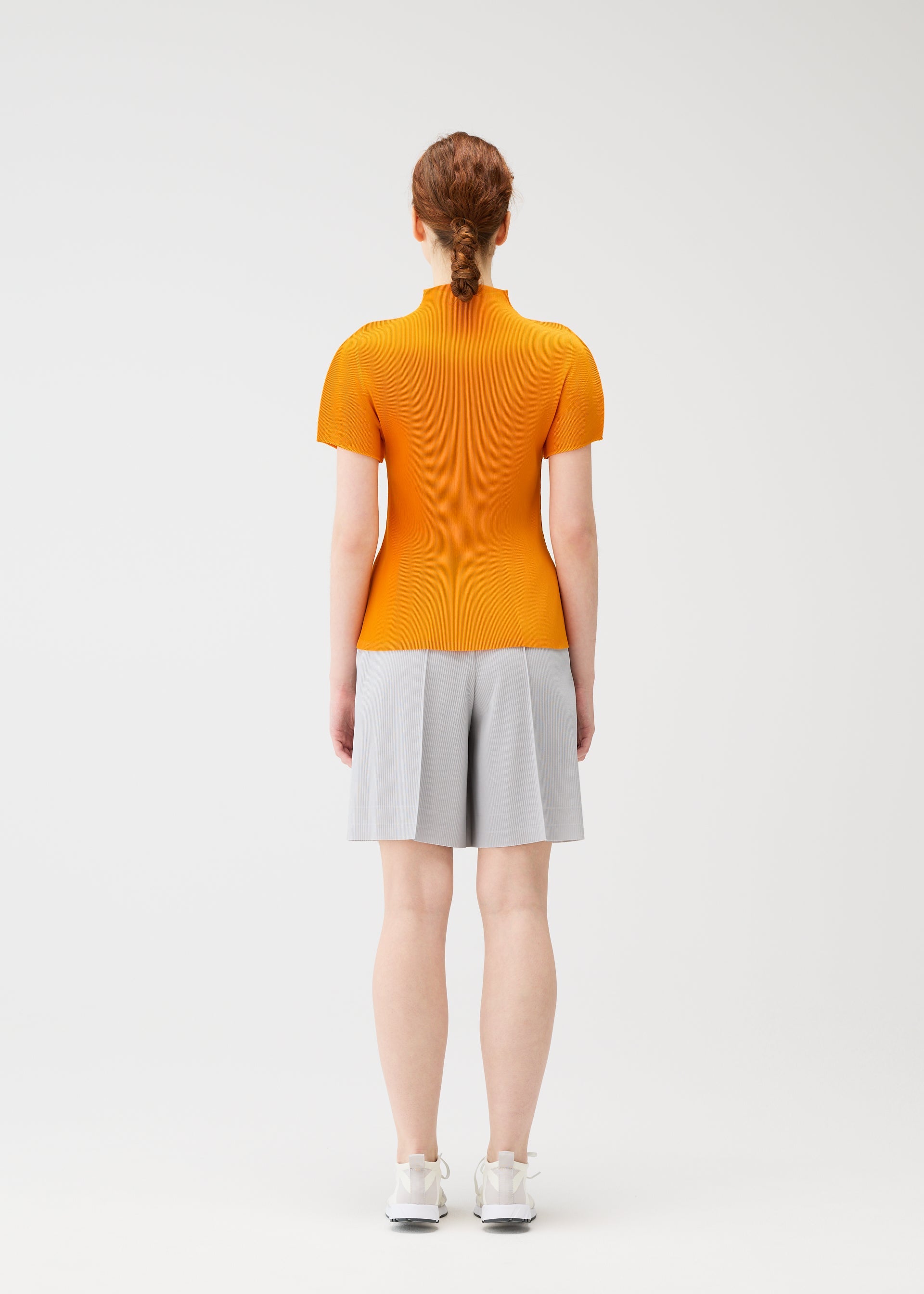 Zoomed view A person with red hair in a low bun stands facing away, wearing an orange short-sleeve top, PLEATS PLEASE ISSEY MIYAKE A-POC BOTTOMS light gray pleated shorts, and white sneakers against a plain white background.