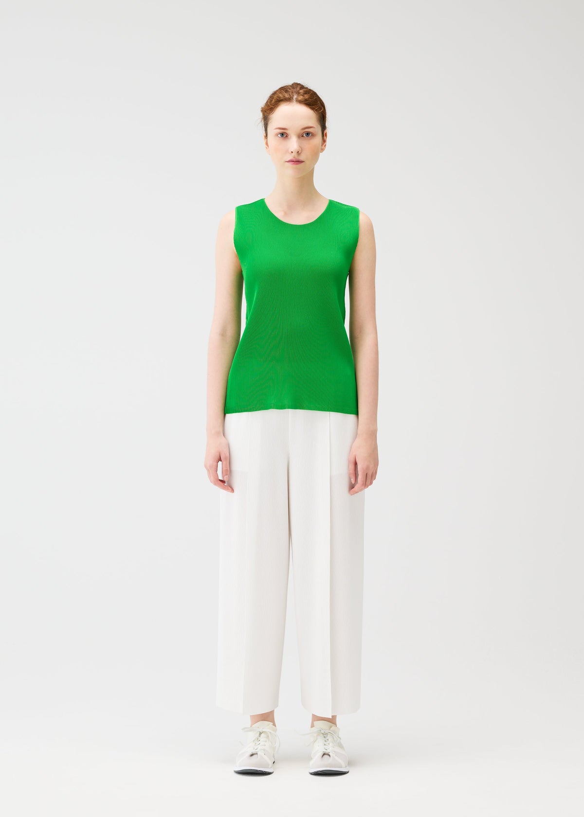 A woman faces forward against a plain white background, wearing PLEATS PLEASE ISSEY MIYAKE's sleeveless green pleated top and A-POC BOTTOMS white wide-leg trousers, paired with white sneakers. view 5 of 8