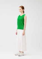 A woman with fair skin and auburn hair in a loose braid stands in a bright studio, facing slightly right. She wears PLEATS PLEASE ISSEY MIYAKE A-POC BOTTOMS—wide pleated pants—paired with a sleeveless green top and white sneakers.