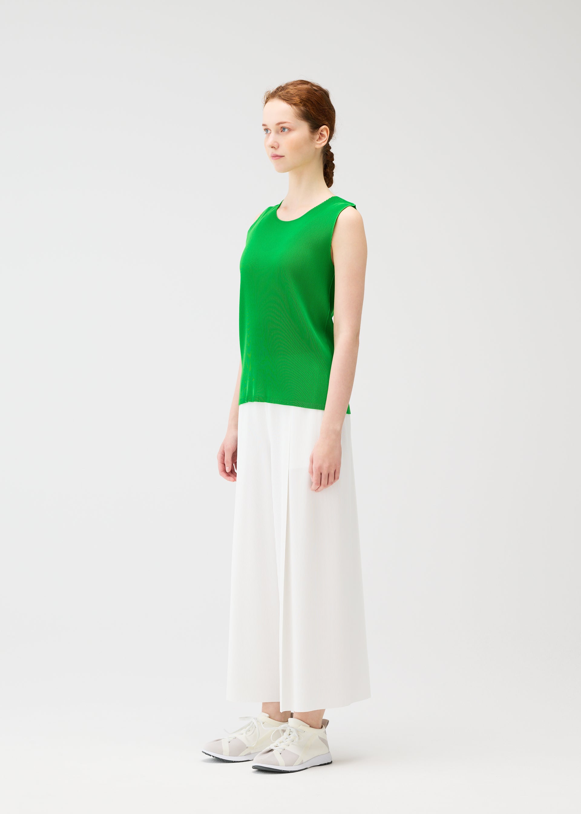 Zoomed view A woman with fair skin and auburn hair in a loose braid stands in a bright studio, facing slightly right. She wears PLEATS PLEASE ISSEY MIYAKE A-POC BOTTOMS—wide pleated pants—paired with a sleeveless green top and white sneakers.
