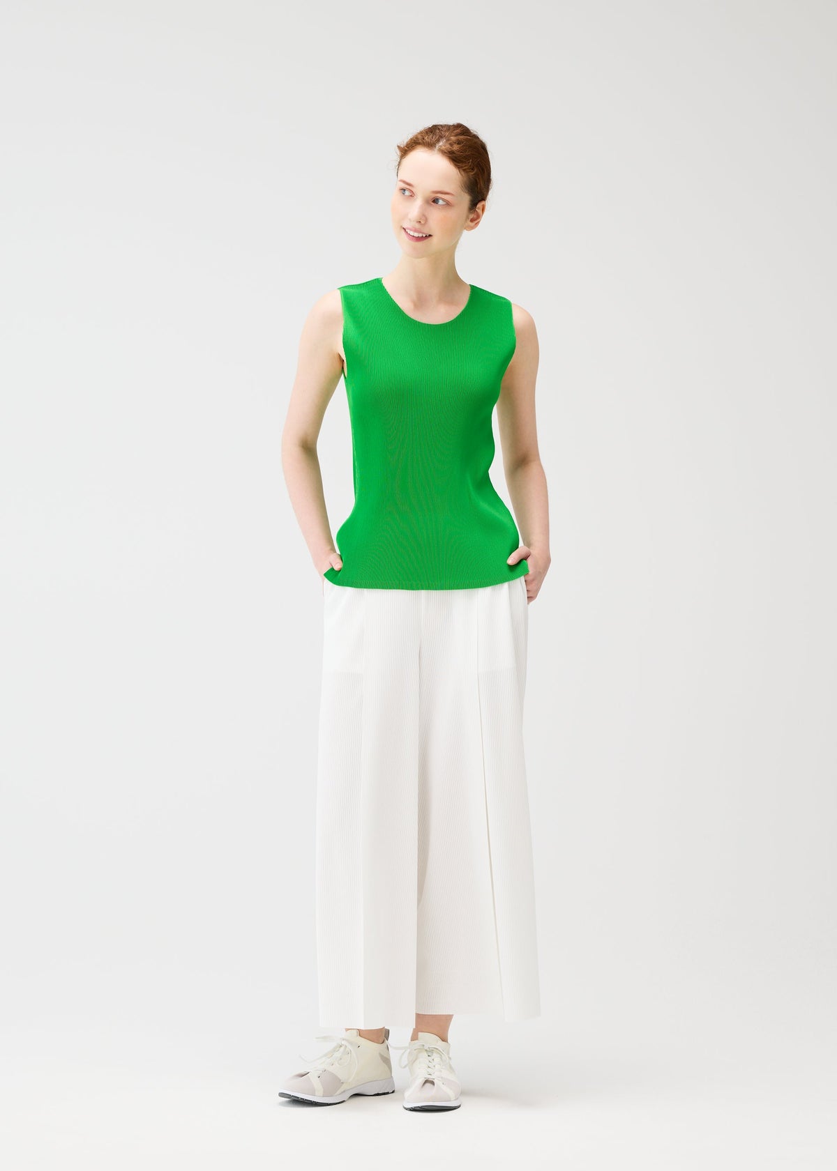 A woman smiles with her hands in her pockets, wearing PLEATS PLEASE ISSEY MIYAKE A-POC BOTTOMS—wide-leg white pleated pants—paired with a sleeveless green top and white sneakers against a plain white background. view 8 of 8