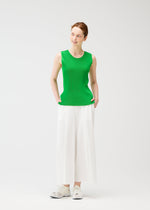 A woman smiles with her hands in her pockets, wearing PLEATS PLEASE ISSEY MIYAKE A-POC BOTTOMS—wide-leg white pleated pants—paired with a sleeveless green top and white sneakers against a plain white background.