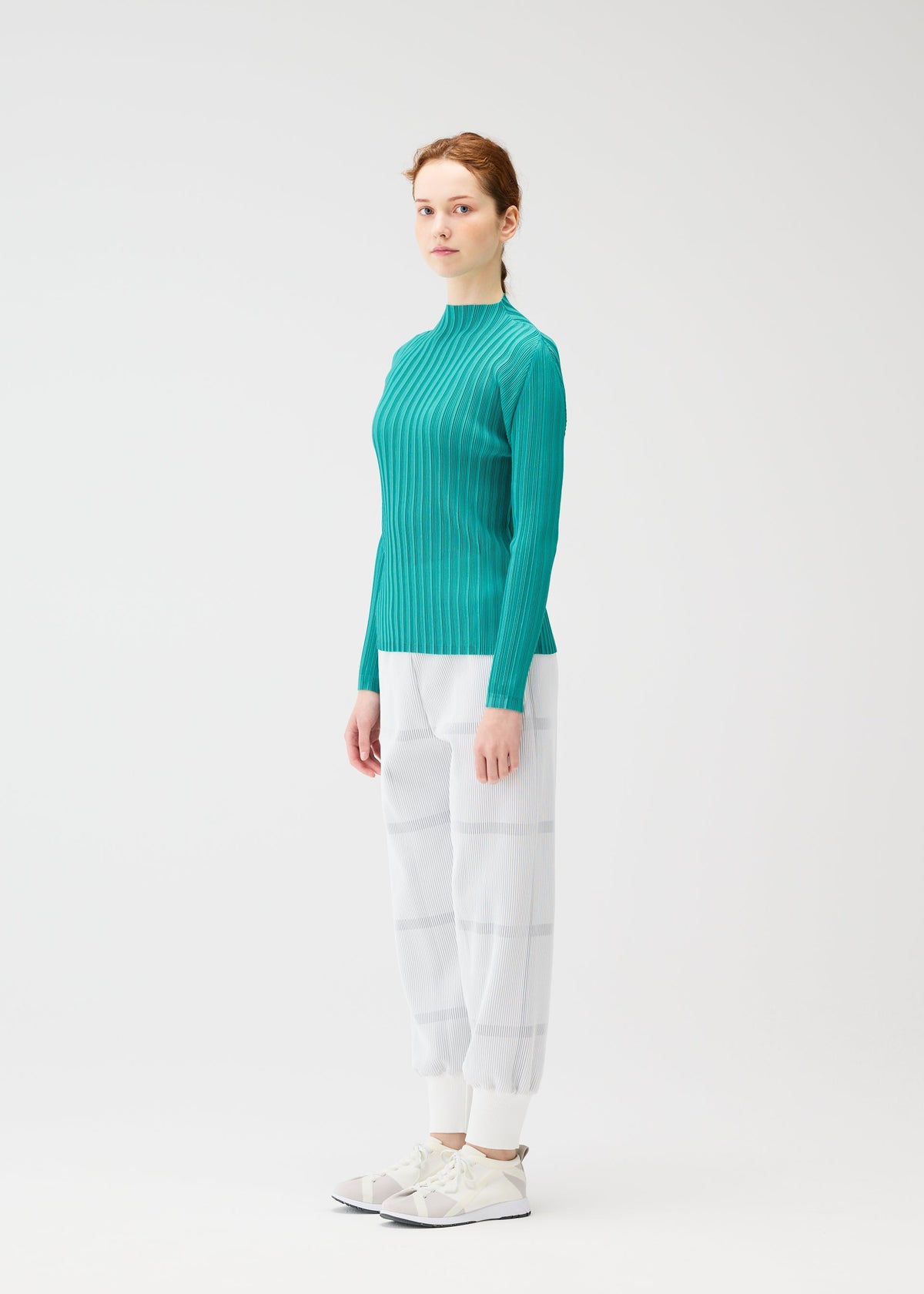 A woman with light skin and auburn hair tied back stands sideways against a plain white background, wearing the PLEATS PLEASE ISSEY MIYAKE CROSSING KNIT in teal, paired with white plaid pants and white sneakers. view 5 of 7