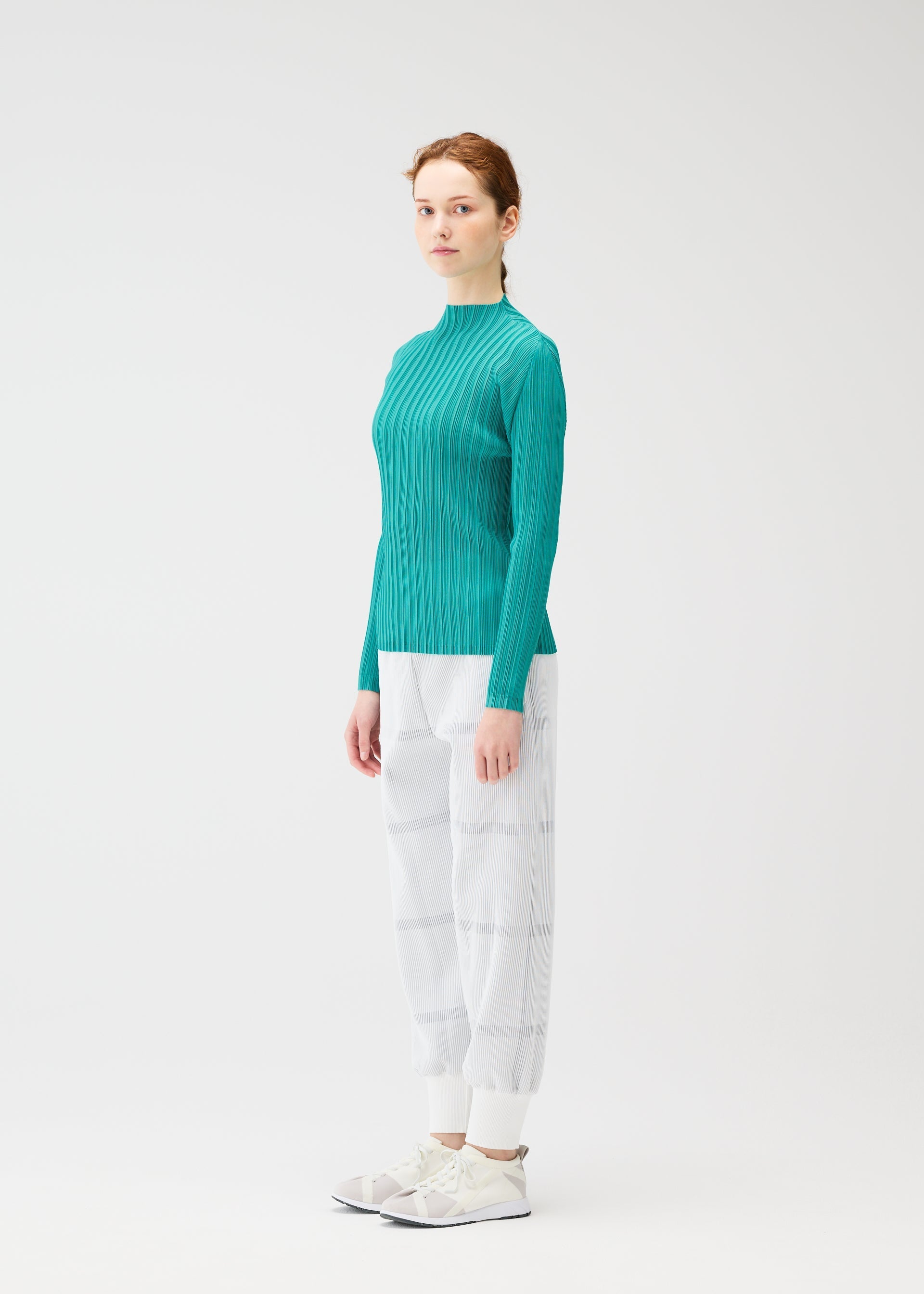 Zoomed view A woman with light skin and auburn hair tied back stands sideways against a plain white background, wearing the PLEATS PLEASE ISSEY MIYAKE CROSSING KNIT in teal, paired with white plaid pants and white sneakers.
