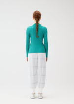 A woman with long, braided red hair faces away, wearing the CROSSING KNIT top by PLEATS PLEASE ISSEY MIYAKE, paired with sheer white wide-leg pants and white shoes against a plain white background.