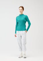 A woman smiles against a white background, wearing the CROSSING KNIT long-sleeve top by PLEATS PLEASE ISSEY MIYAKE in teal ribbed knit, paired with light gray plaid pants and white sneakers. Her right hand is in her pocket.