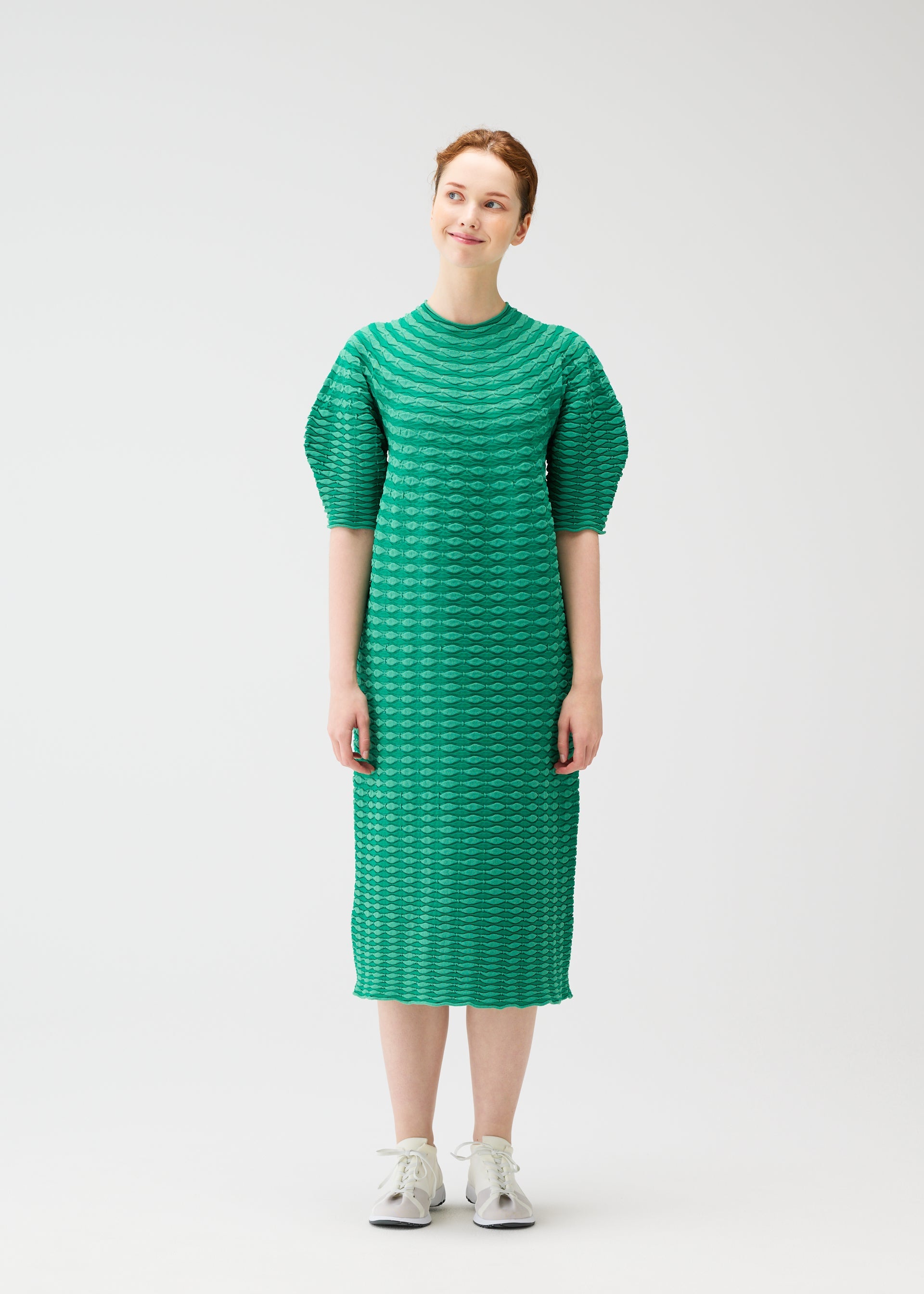 Zoomed view A woman wears the COLORFUL MUSHROOM KNIT by PLEATS PLEASE ISSEY MIYAKE, a textured green knee-length mock-neck dress with short puff sleeves. She pairs it with light sneakers and smiles gently; the dress is machine washable for easy care.