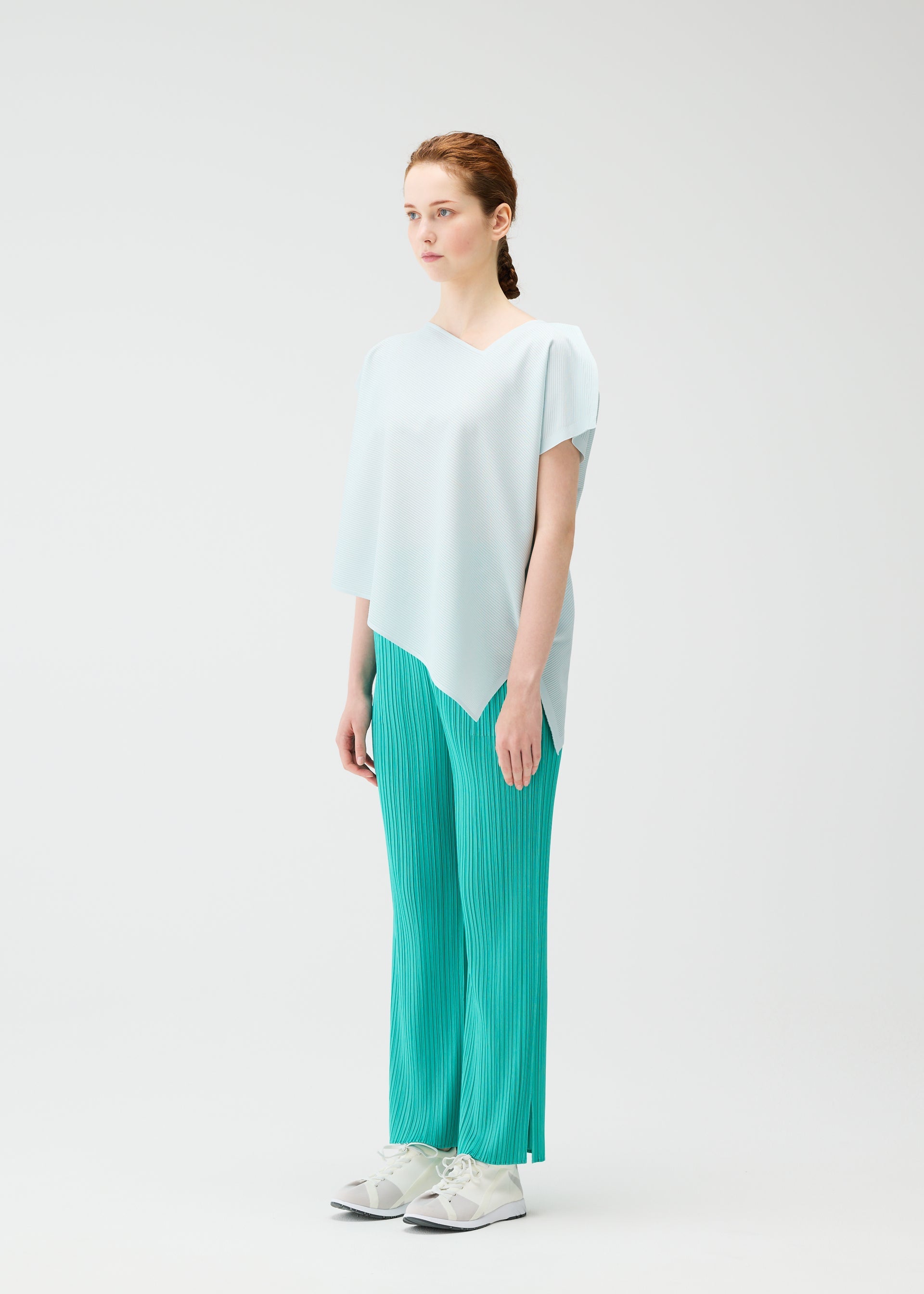 Zoomed view A woman with light skin and red hair stands against a plain white background, wearing the PLEATS PLEASE ISSEY MIYAKE A-POC SEA GLASS top, teal pleated pants, and white sneakers. She looks slightly to the side with a neutral expression.