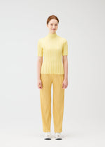 A woman faces forward, smiling softly, dressed in the PLEATS PLEASE ISSEY MIYAKE RIB KNIT light yellow short-sleeved turtleneck top and matching pleated pants with bold lines, set against a plain white background.