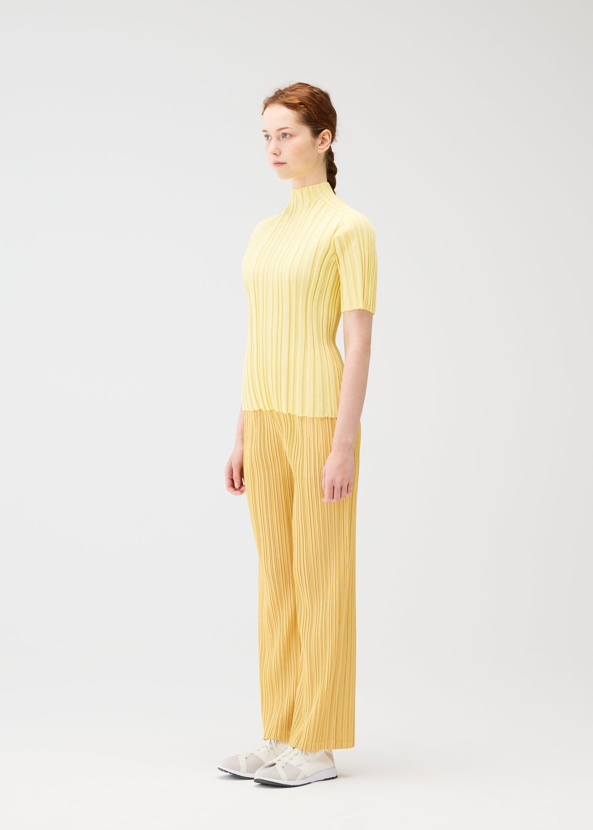 A light-skinned, reddish-haired person in profile stands against a white background wearing the PLEATS PLEASE ISSEY MIYAKE RIB KNIT yellow top and mustard yellow pleated pants with white sneakers. view 6 of 7