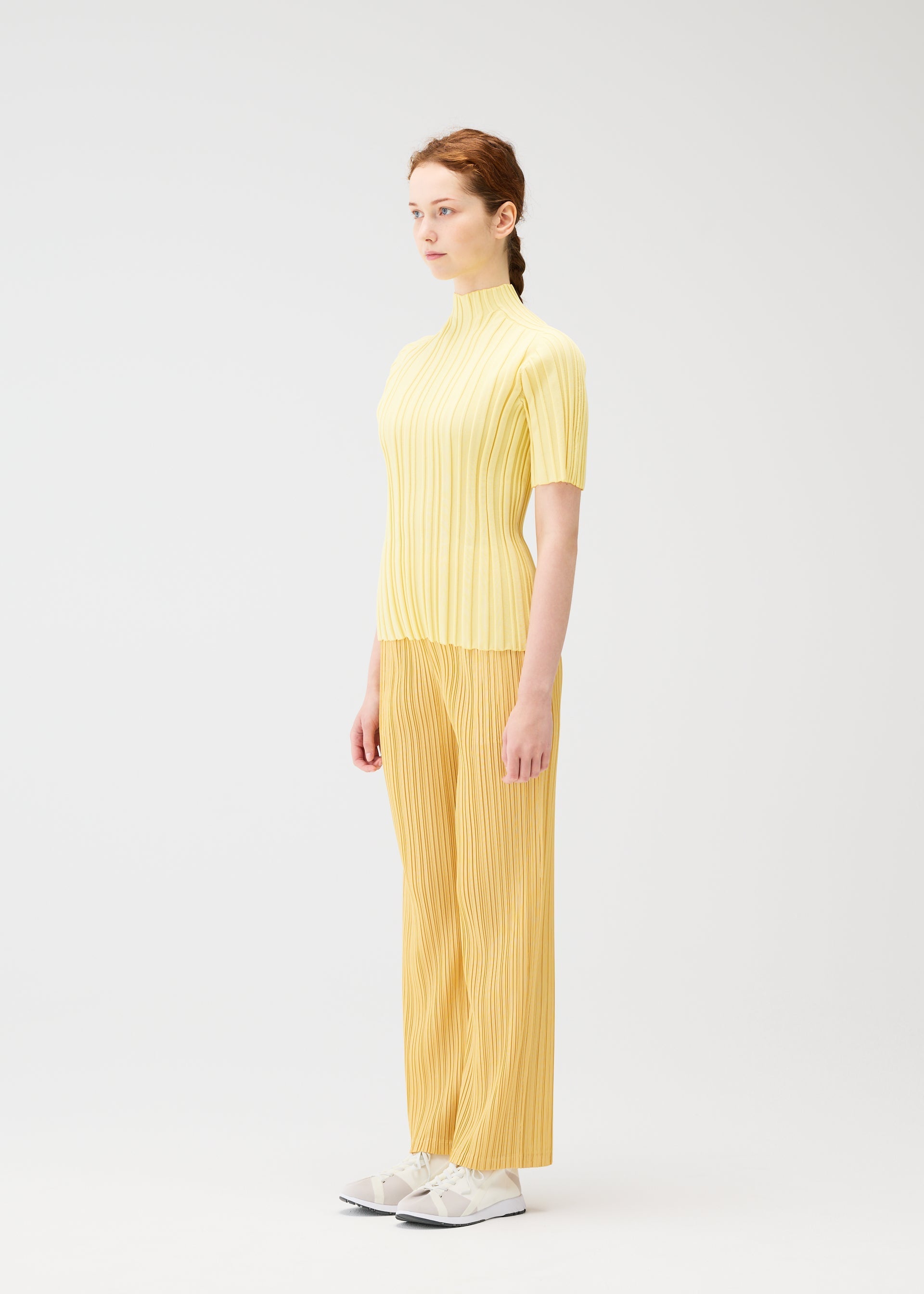 Zoomed view A light-skinned, reddish-haired person in profile stands against a white background wearing the PLEATS PLEASE ISSEY MIYAKE RIB KNIT yellow top and mustard yellow pleated pants with white sneakers.
