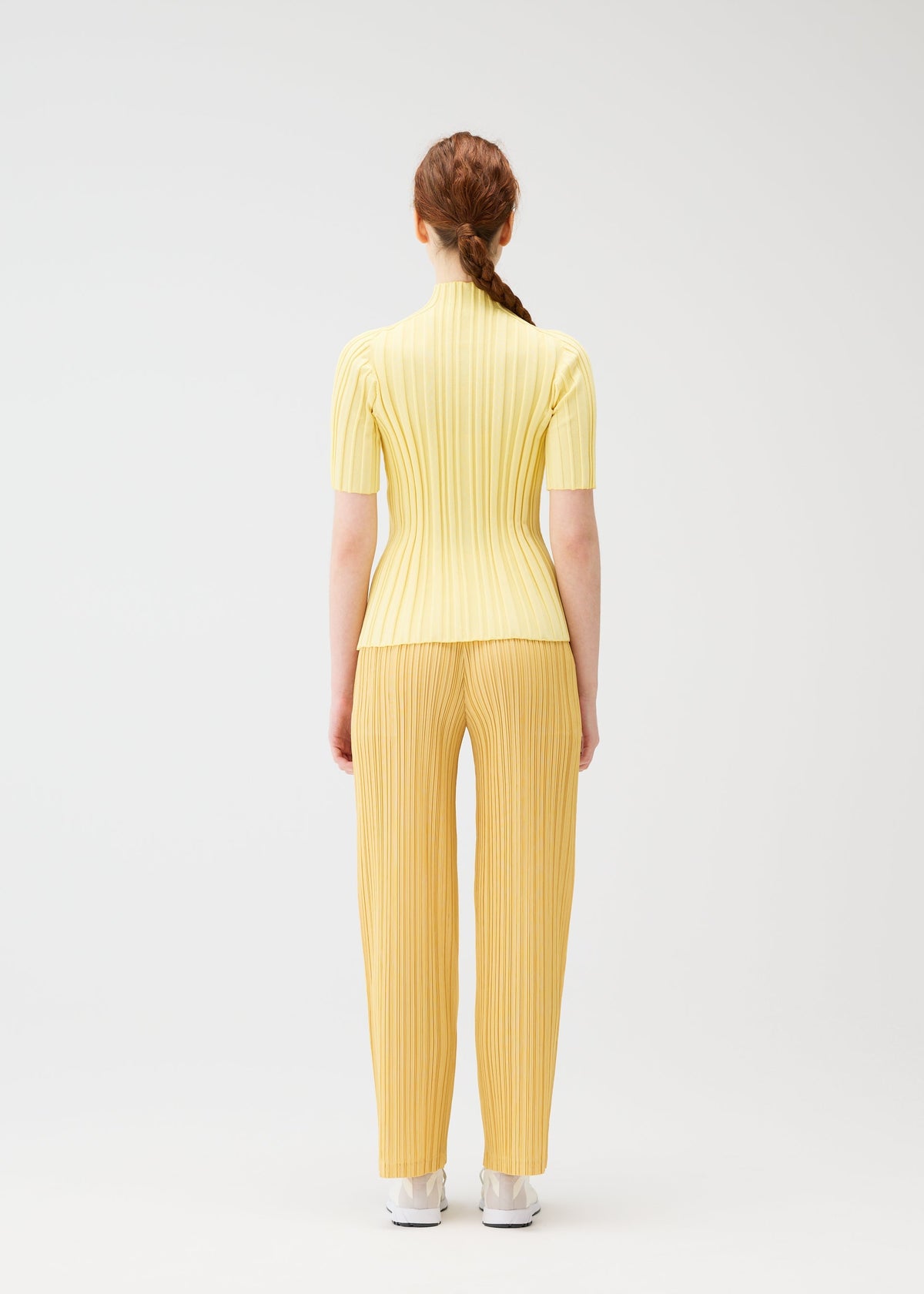A person with long red hair in a braid stands facing away, wearing the yellow RIB KNIT set from PLEATS PLEASE ISSEY MIYAKE and white shoes, against a plain white background. view 7 of 7
