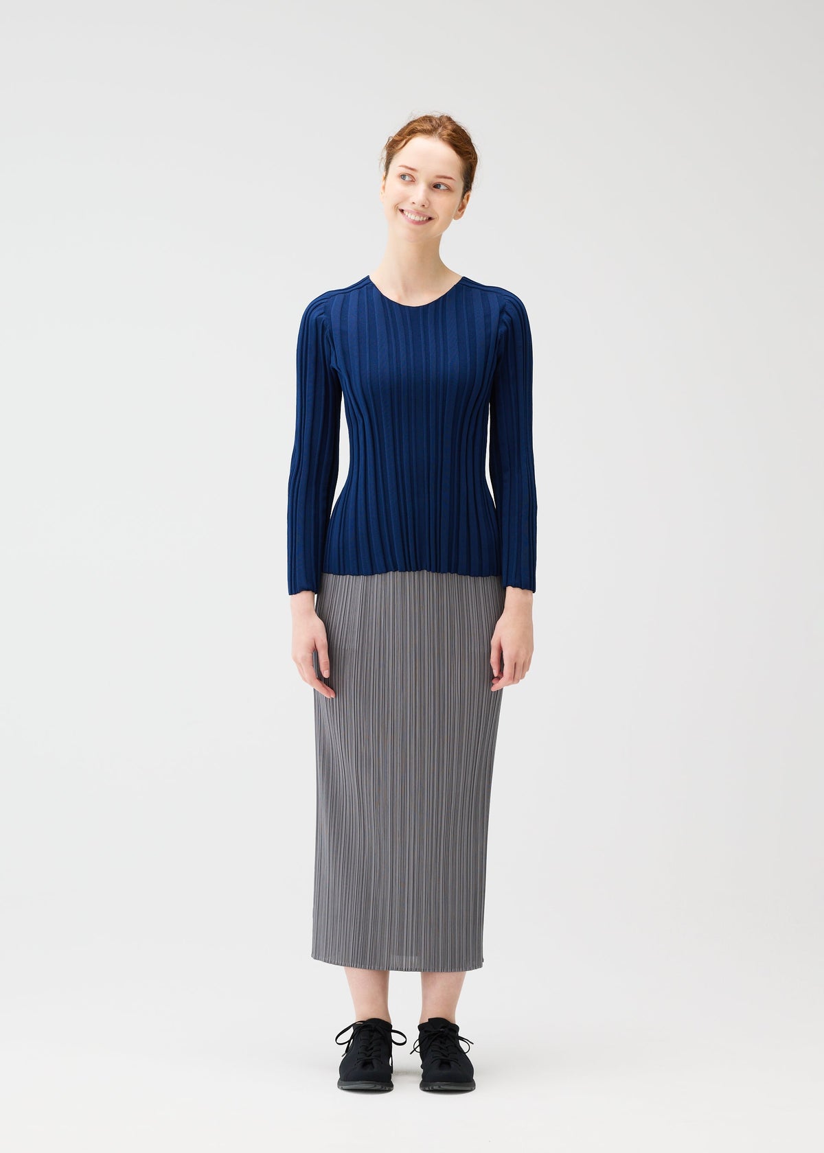 A woman with red hair tied back stands smiling against a white background, wearing the PLEATS PLEASE ISSEY MIYAKE RIB KNIT navy long-sleeved top, a gray pleated midi skirt, and black shoes. view 4 of 7