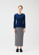 A woman with red hair tied back stands smiling against a white background, wearing the PLEATS PLEASE ISSEY MIYAKE RIB KNIT navy long-sleeved top, a gray pleated midi skirt, and black shoes.