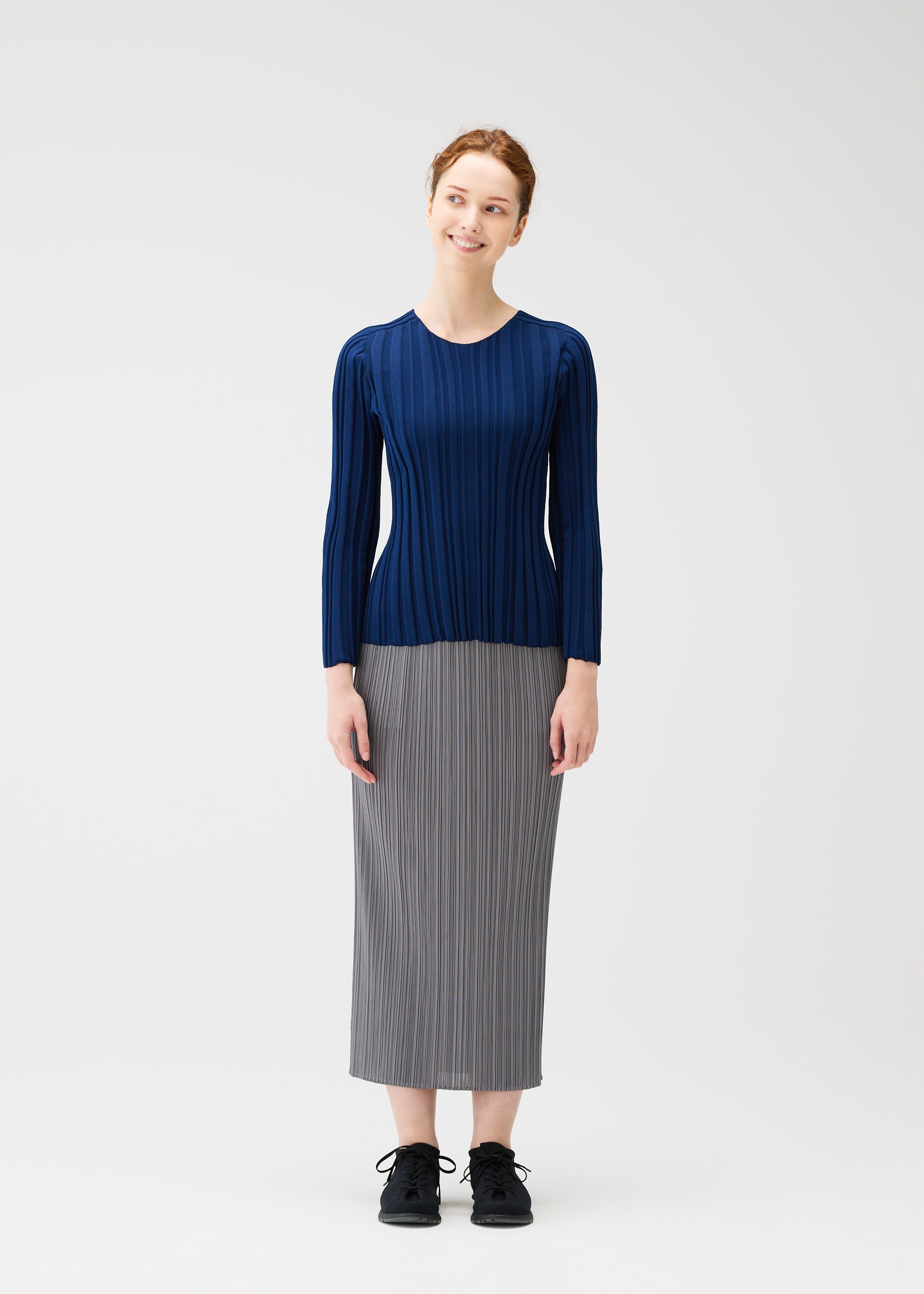 Zoomed view A woman with red hair tied back stands smiling against a white background, wearing the PLEATS PLEASE ISSEY MIYAKE RIB KNIT navy long-sleeved top, a gray pleated midi skirt, and black shoes.