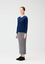A woman with light skin and reddish hair tied back smiles, wearing a navy blue RIB KNIT by PLEATS PLEASE ISSEY MIYAKE, a gray pleated skirt, and black shoes against a plain white background.