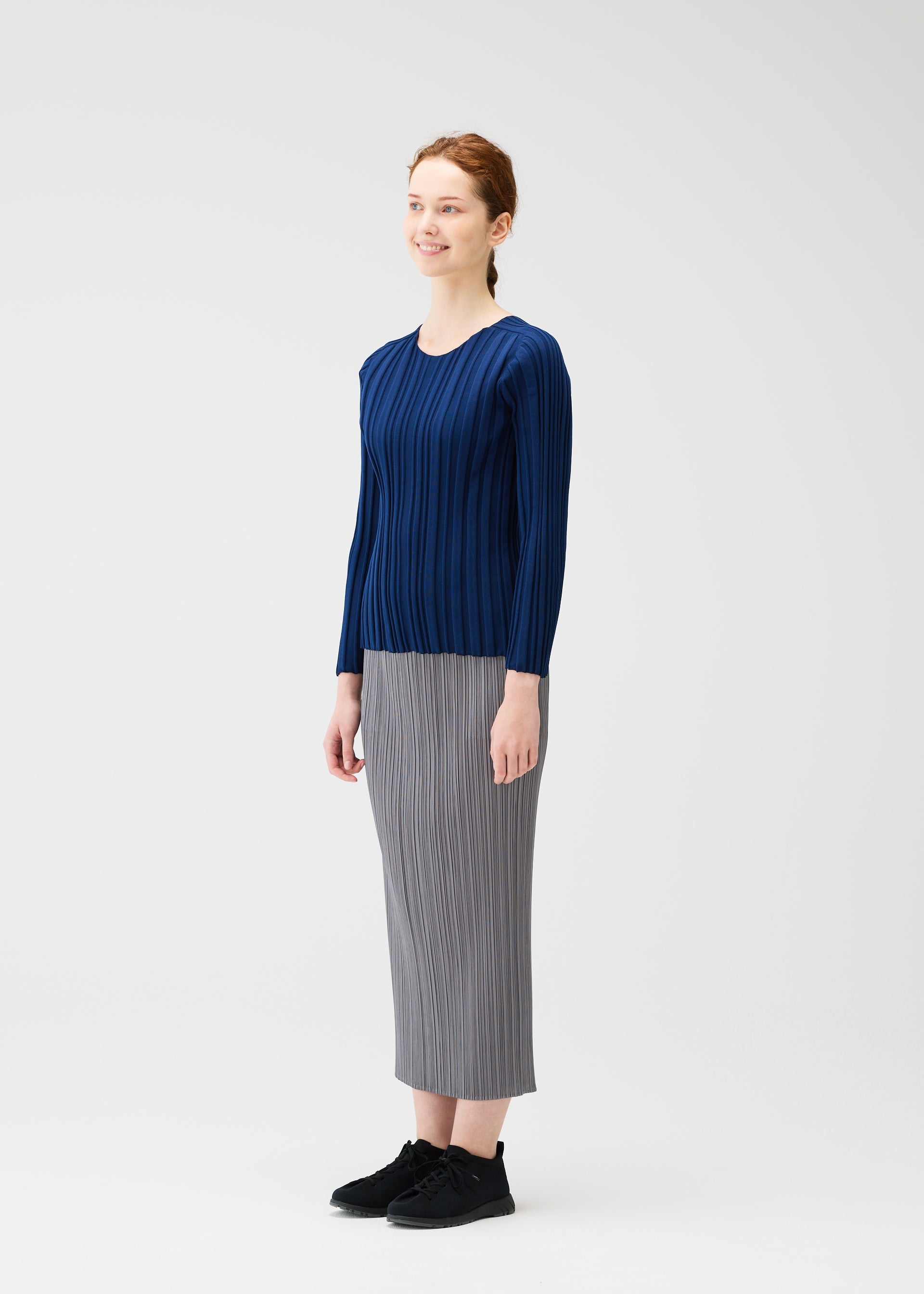 Zoomed view A woman with light skin and reddish hair tied back smiles, wearing a navy blue RIB KNIT by PLEATS PLEASE ISSEY MIYAKE, a gray pleated skirt, and black shoes against a plain white background.