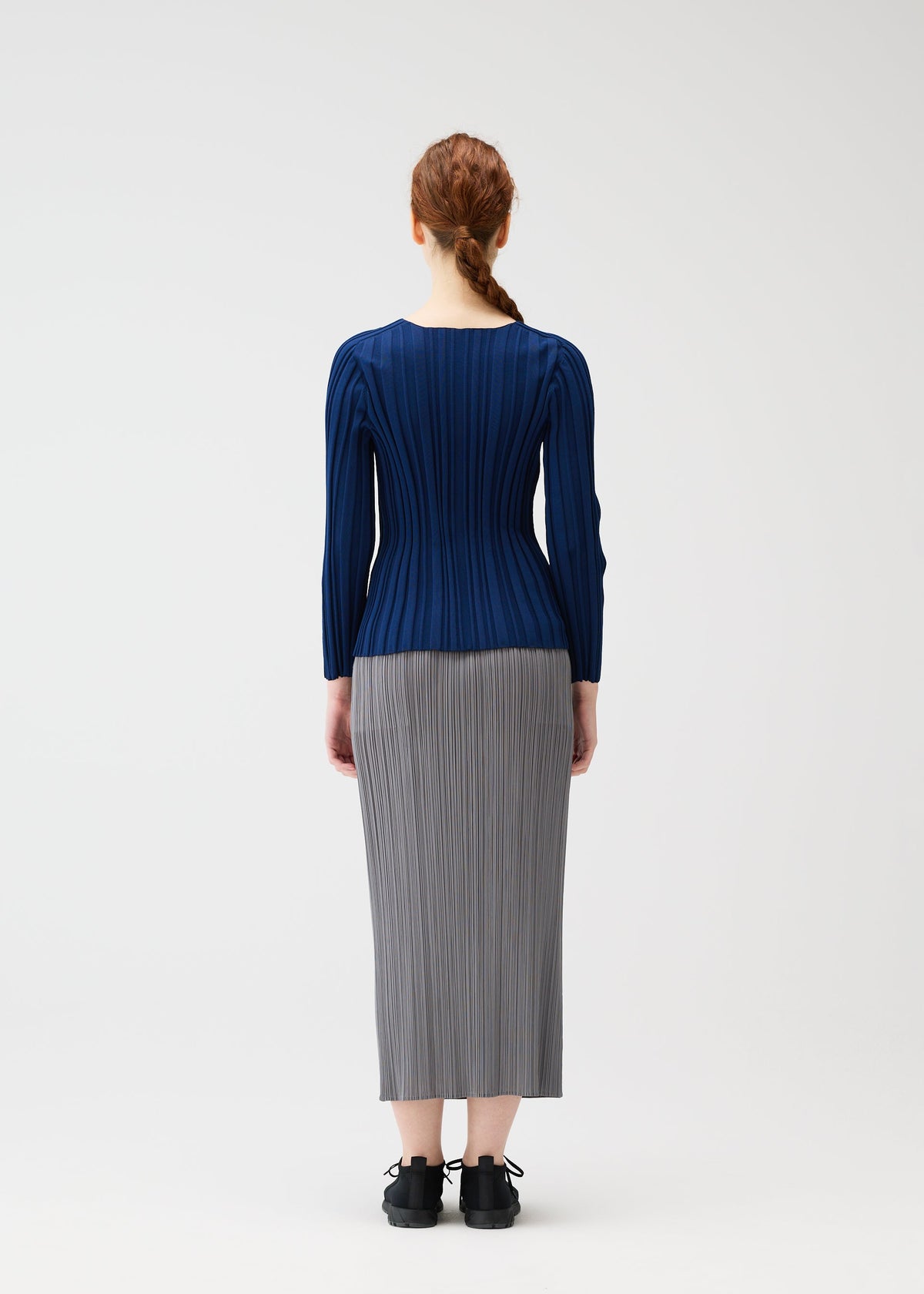 A person with long red hair in a braid faces away, wearing a fitted blue RIB KNIT top and gray pleated skirt by PLEATS PLEASE ISSEY MIYAKE, with black shoes, against a plain light background. view 6 of 7