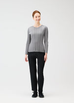 A woman smiles against a plain white background, wearing the PLEATS PLEASE ISSEY MIYAKE RIB KNIT top paired with black pleated pants and black shoes.