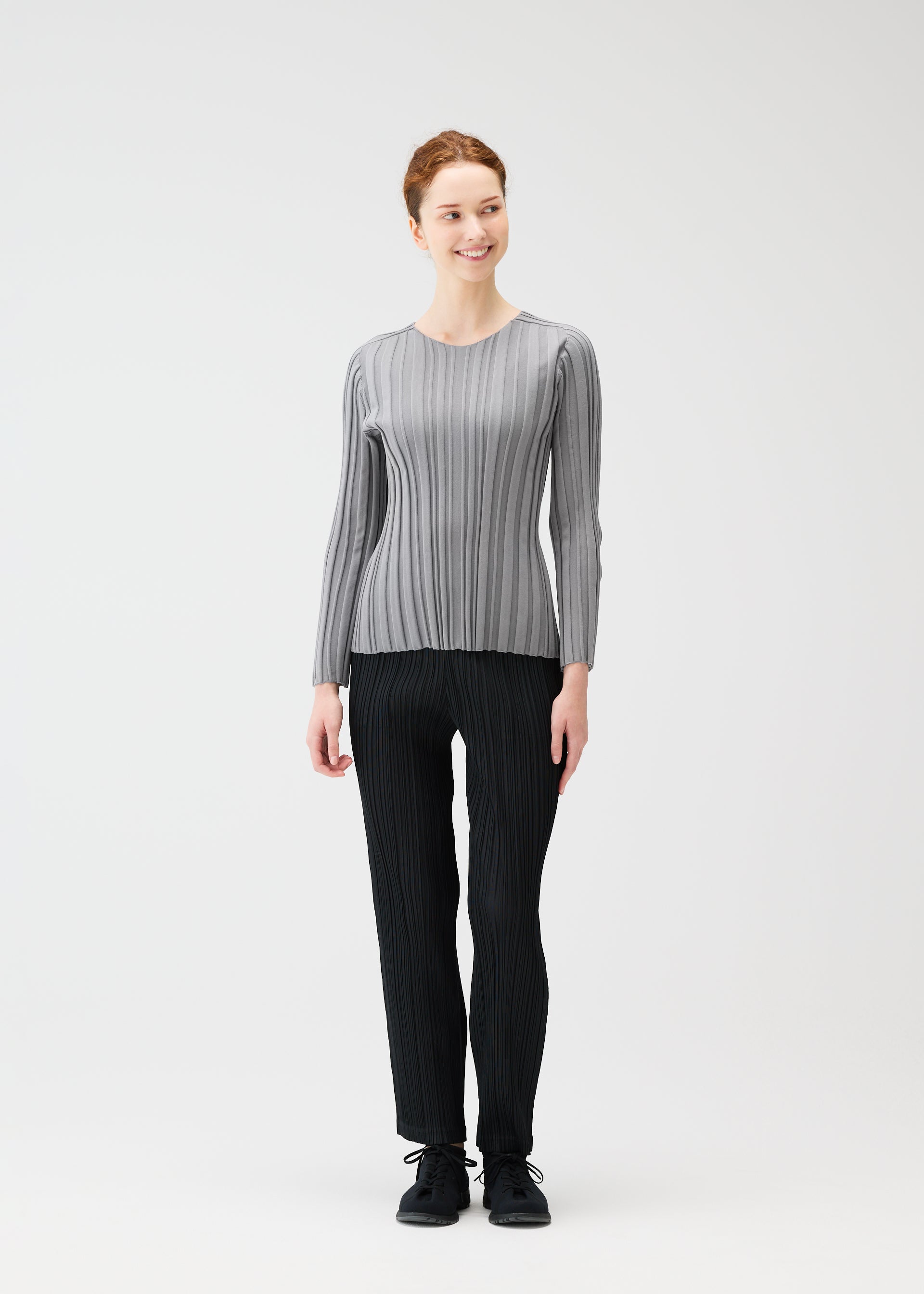 Zoomed view A woman smiles against a plain white background, wearing the PLEATS PLEASE ISSEY MIYAKE RIB KNIT top paired with black pleated pants and black shoes.