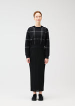 A woman stands facing forward, smiling slightly with her hair tied back. She wears the PLEATS PLEASE ISSEY MIYAKE CROSSING KNIT city-inspired round-neck sweater, a long black pleated skirt, and black sneakers against a plain white background.