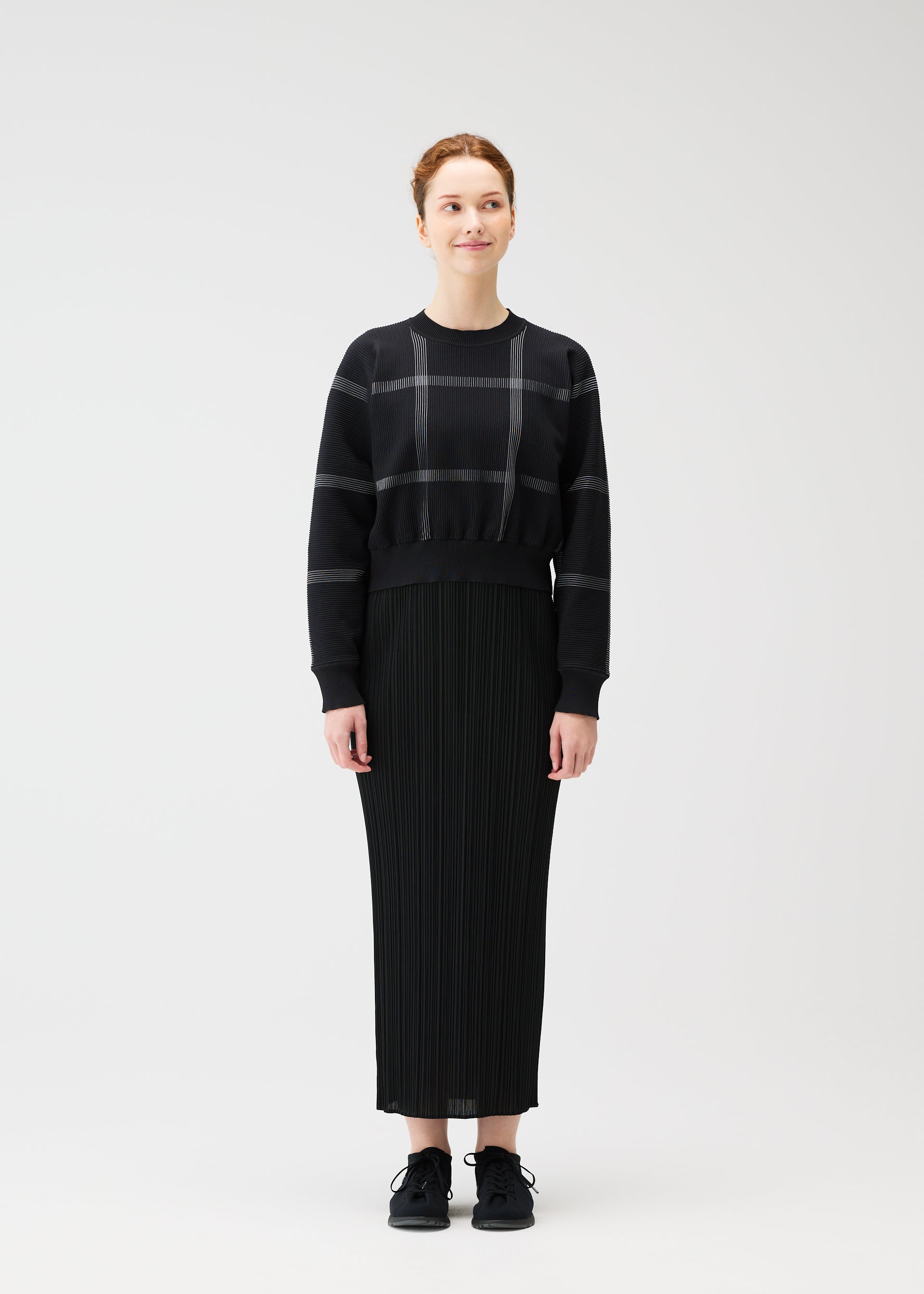 Zoomed view A woman stands facing forward, smiling slightly with her hair tied back. She wears the PLEATS PLEASE ISSEY MIYAKE CROSSING KNIT city-inspired round-neck sweater, a long black pleated skirt, and black sneakers against a plain white background.