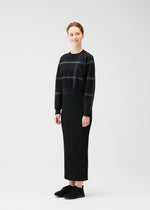 A person smiles and looks to the side, wearing the CROSSING KNIT sweater by PLEATS PLEASE ISSEY MIYAKE with subtle stripes, a long ribbed black skirt, and black sneakers in a studio setting.