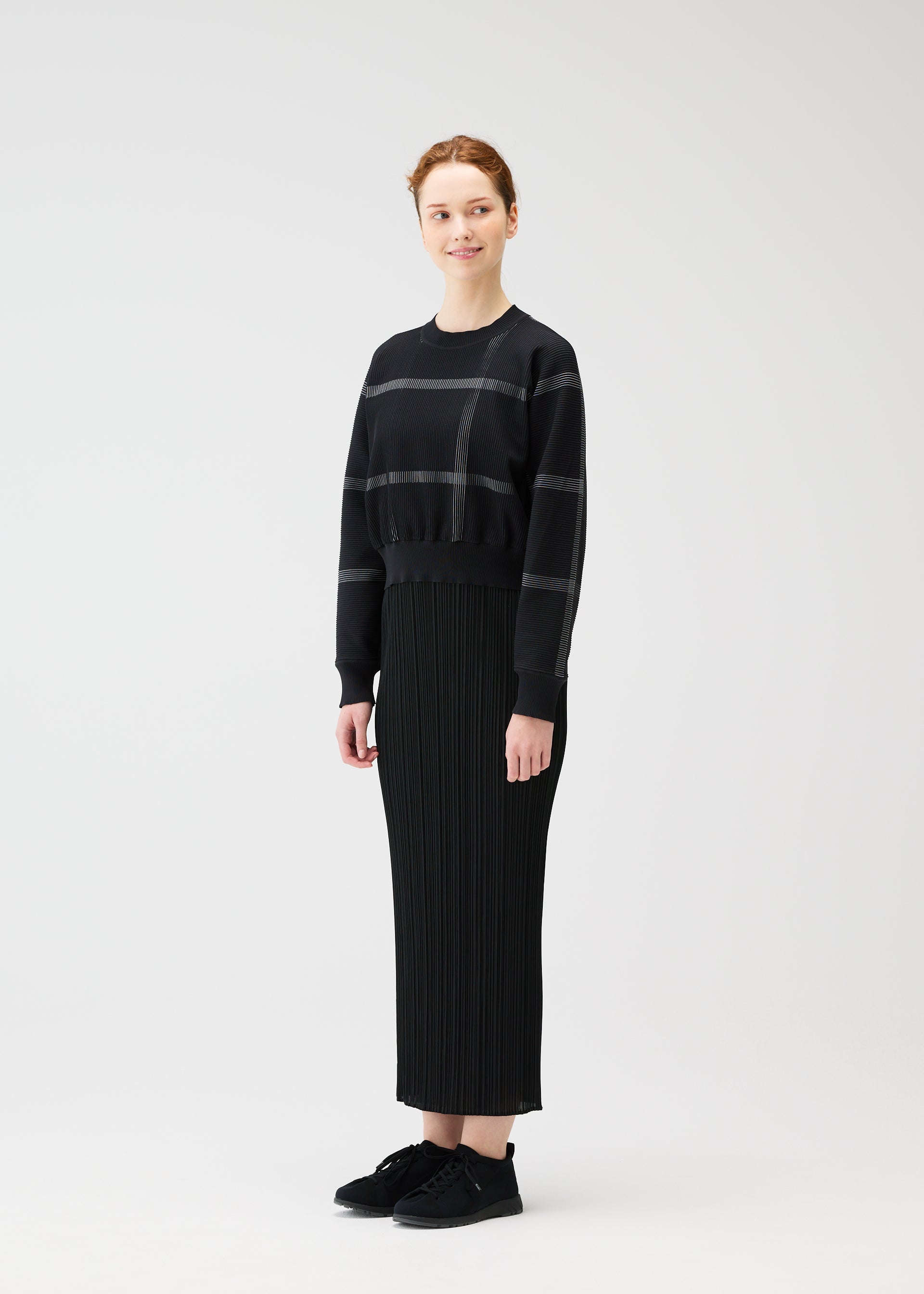 Zoomed view A person smiles and looks to the side, wearing the CROSSING KNIT sweater by PLEATS PLEASE ISSEY MIYAKE with subtle stripes, a long ribbed black skirt, and black sneakers in a studio setting.