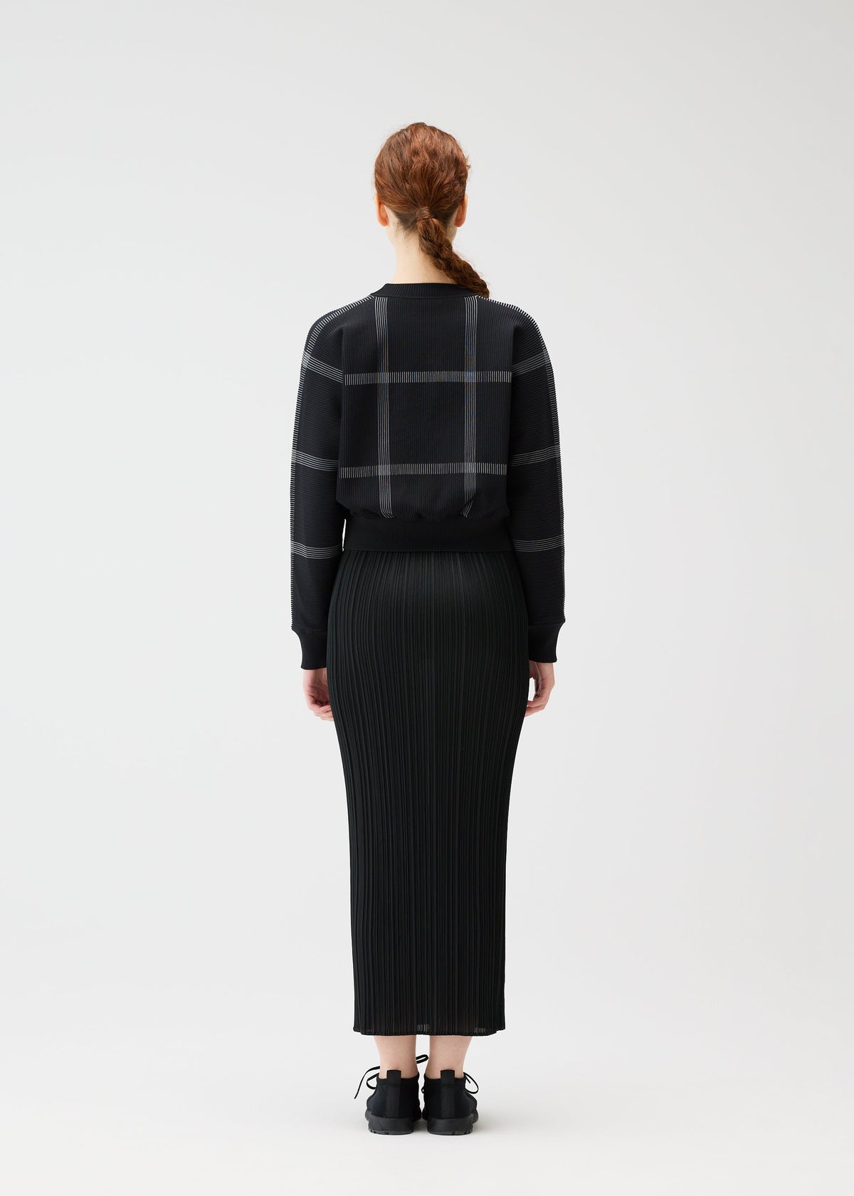 A person with long, reddish-brown braided hair stands facing away, wearing the PLEATS PLEASE ISSEY MIYAKE CROSSING KNIT sweater with a city-inspired plaid pattern and a long black pleated skirt, set against a plain background. view 6 of 6