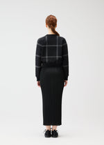 A person with long, reddish-brown braided hair stands facing away, wearing the PLEATS PLEASE ISSEY MIYAKE CROSSING KNIT sweater with a city-inspired plaid pattern and a long black pleated skirt, set against a plain background.