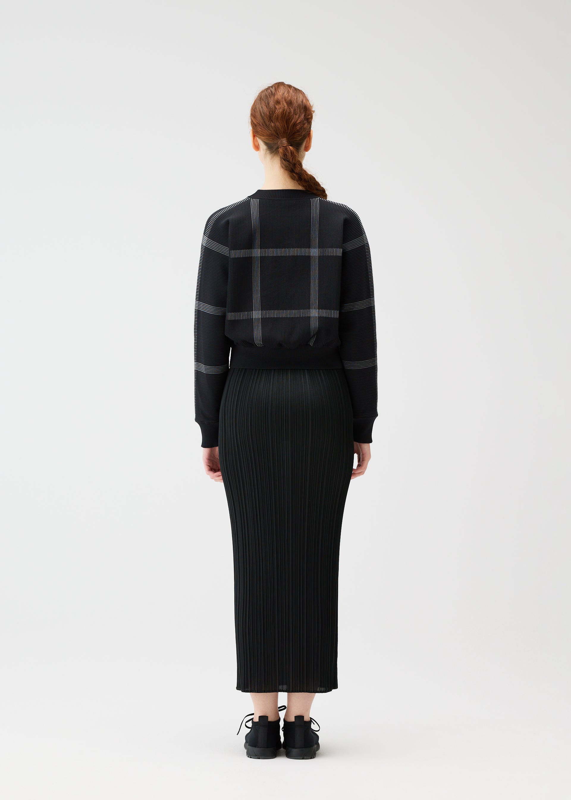 Zoomed view A person with long, reddish-brown braided hair stands facing away, wearing the PLEATS PLEASE ISSEY MIYAKE CROSSING KNIT sweater with a city-inspired plaid pattern and a long black pleated skirt, set against a plain background.