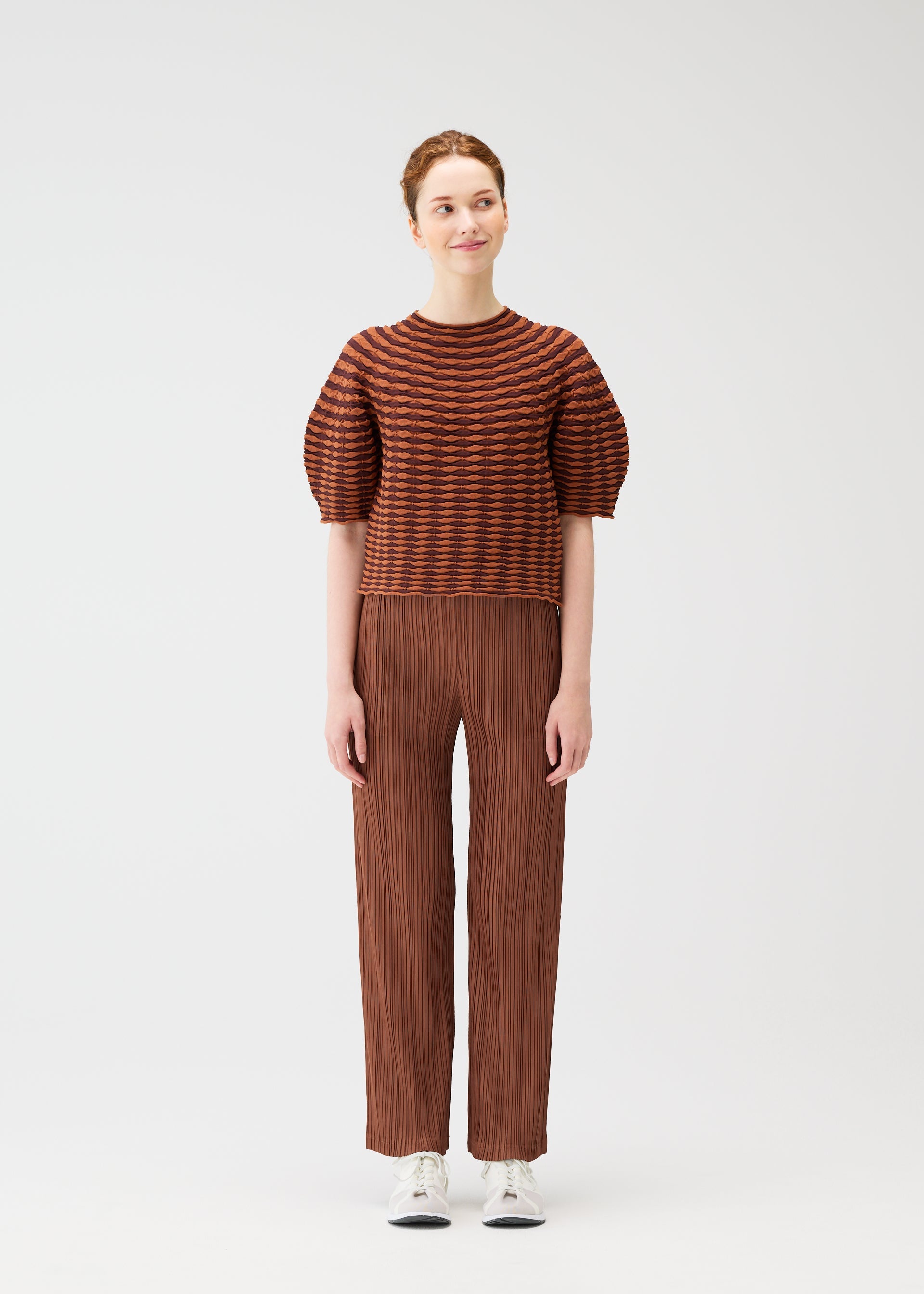 Zoomed view A person with light skin smiles slightly while looking to the side, wearing the PLEATS PLEASE ISSEY MIYAKE COLORFUL MUSHROOM KNIT top, brown pleated pants, and white sneakers. The machine washable outfit is shown against a plain white background.