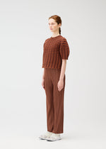 A woman stands in profile against a white background, wearing the COLORFUL MUSHROOM KNIT by PLEATS PLEASE ISSEY MIYAKE with brown pleated pants and white sneakers. Her hair is tied back in a braid.