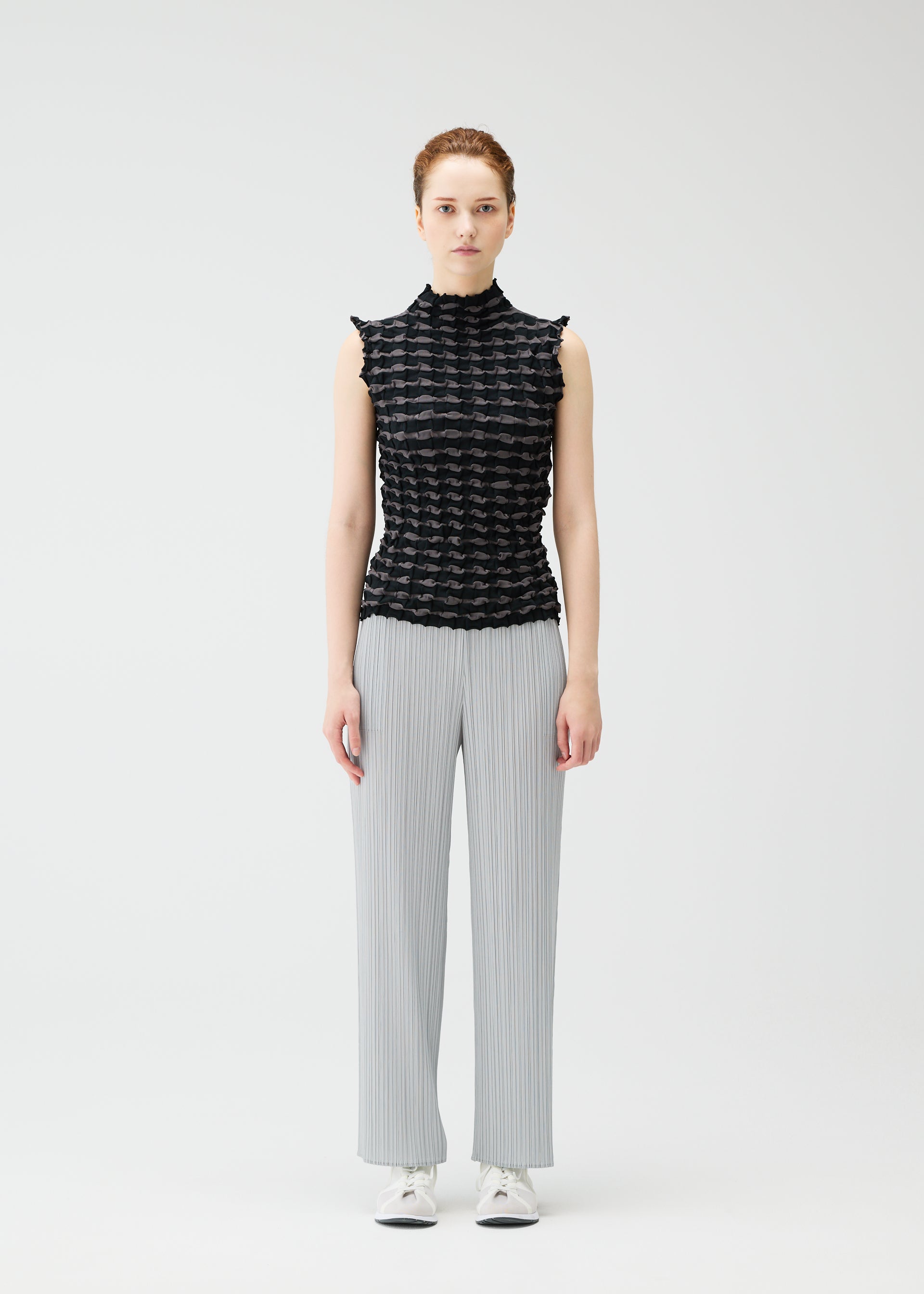 Zoomed view A person with light skin and brown hair tied back stands against a plain white background, wearing the PLEATS PLEASE ISSEY MIYAKE TWIST KNIT, light gray pleated wide-leg pants, and white sneakers.