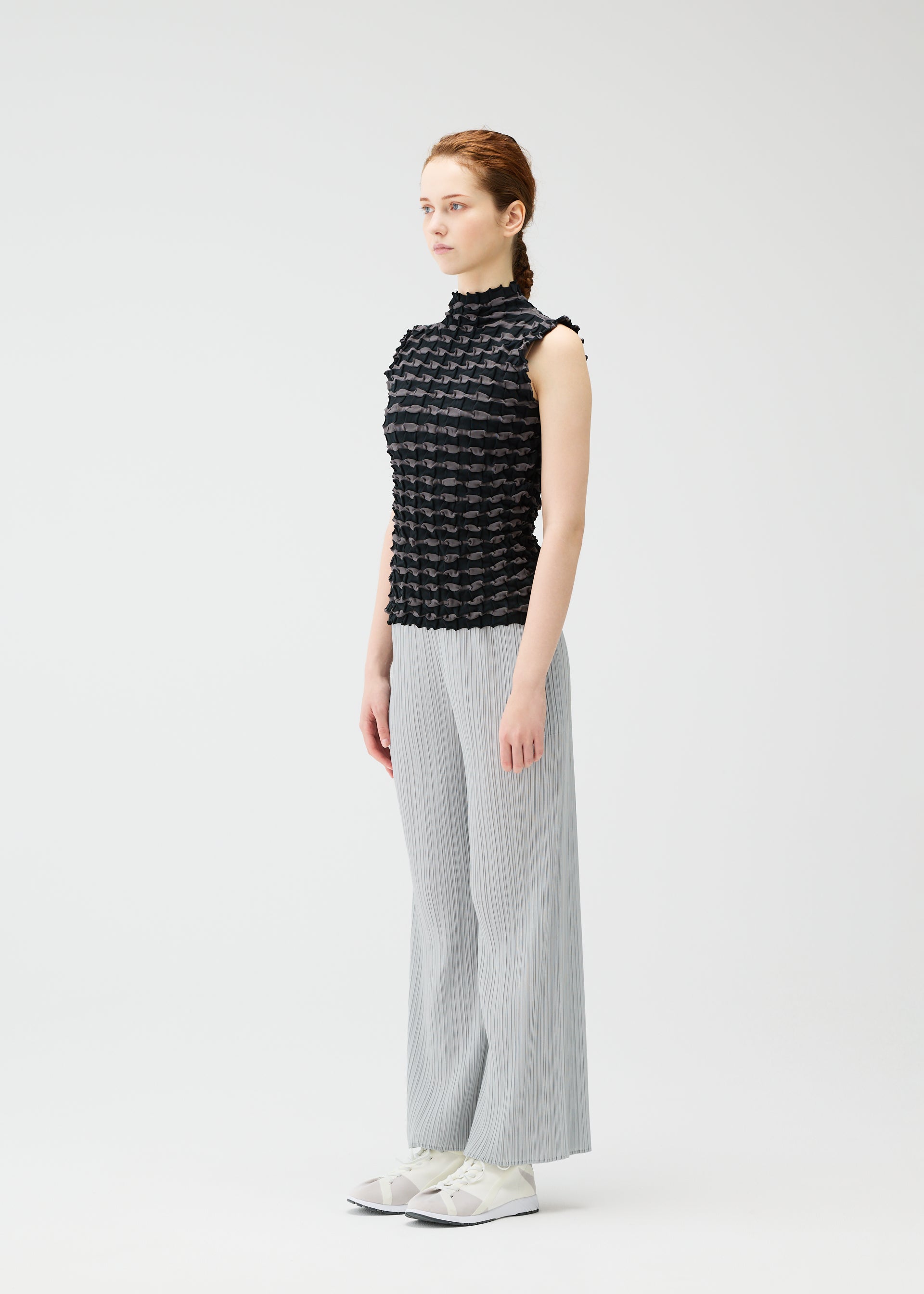 Zoomed view A woman in profile stands against a plain white background, wearing the PLEATS PLEASE ISSEY MIYAKE TWIST KNIT top with light gray wide-leg pants and white sneakers, her hair neatly tied back in a braid.