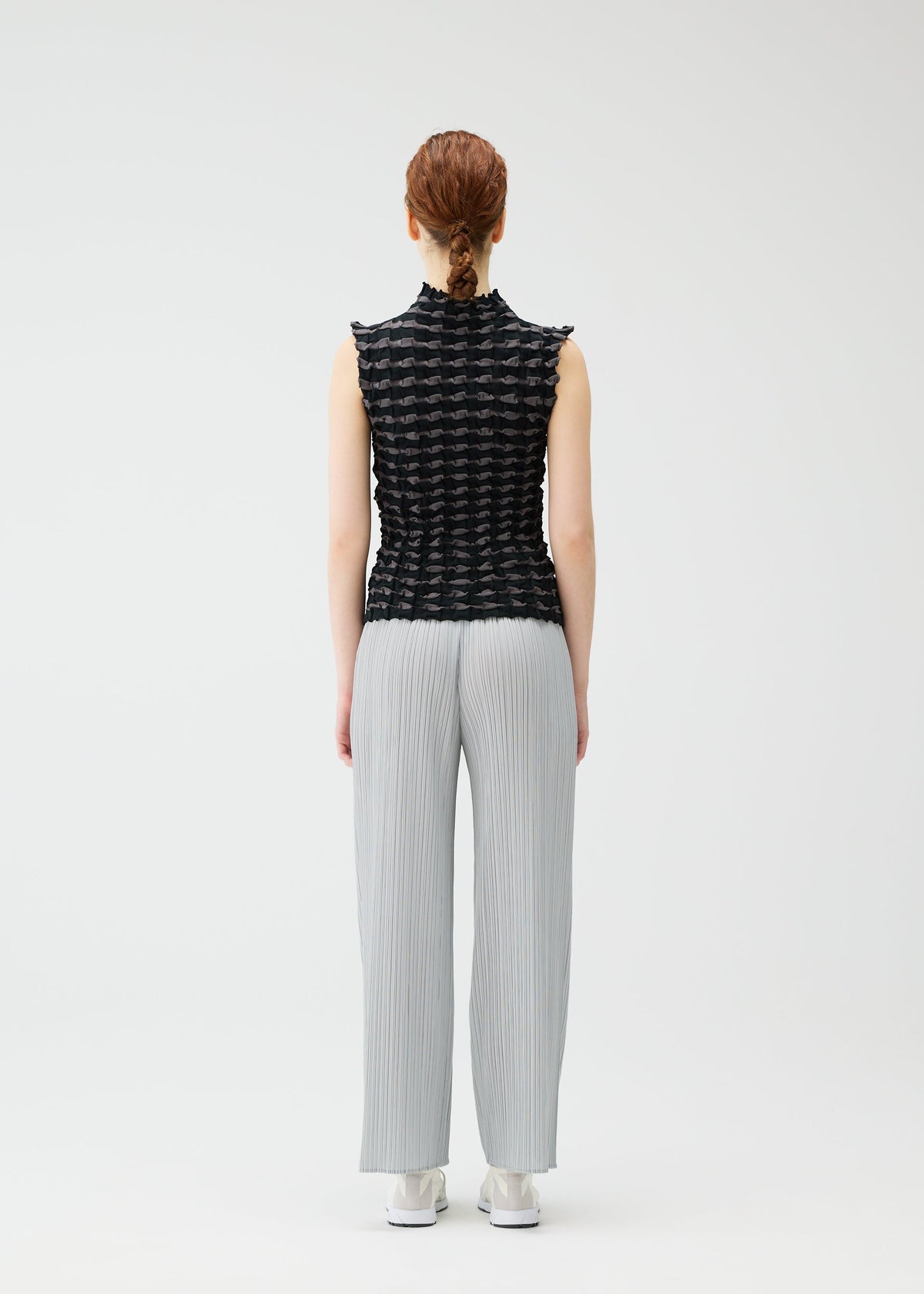 A person with reddish braided hair stands facing away, wearing the PLEATS PLEASE ISSEY MIYAKE TWIST KNIT top with light gray pleated pants and white shoes against a plain white background. view 7 of 8