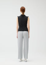 A person with reddish braided hair stands facing away, wearing the PLEATS PLEASE ISSEY MIYAKE TWIST KNIT top with light gray pleated pants and white shoes against a plain white background.